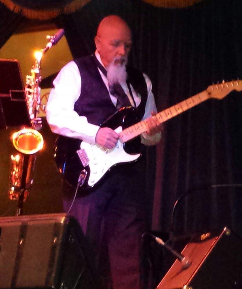 Raul playing electric guitar on stage with his saxophone beside him